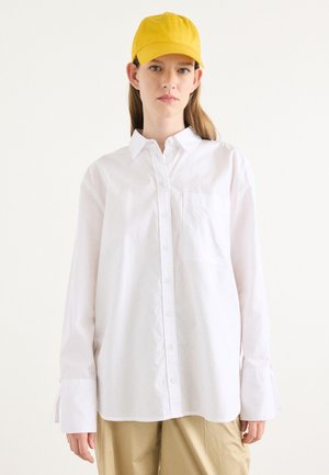 White long-sleeve shirt with a button-down collar and a pocket, paired with yellow cap. Fabric appears smooth; sleeves have a slight flare.