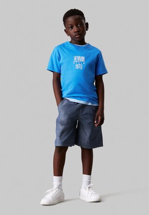 Lichtblauw katoenen t-shirt met witte tekst, gecombineerd met donkerblauwe shorts. Witte sneakers en geribde witte sokken maken de outfit compleet.
