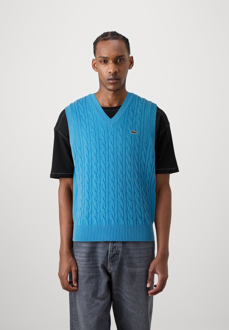 Gilet en tricot à col en V de couleur bleu vif avec un motif de côtes texturé, présentant un ourlet en côtes. Porté sur une chemise noire à manches courtes.