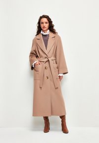 Abrigo beige abierto con mangas anchas, dos bolsillos frontales y un cinturón. Presenta grandes botones dorados y una prenda texturizada con volantes.