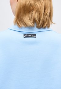 Ξανθό άτομο με μαλλιά μέχρι τους ώμους που φοράει ανοιχτό μπλε πουκάμισο με γιακά και μαύρη ετικέτα "Lacoste" στο πίσω μέρος του γιακά.