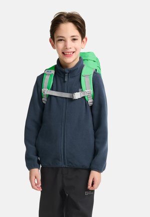 Ragazzo che indossa una giacca in pile blu navy e pantaloni neri con uno zaino verde brillante, sorridente su uno sfondo bianco.