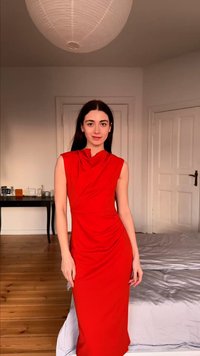 Femme aux longs cheveux foncés portant une robe rouge sans manches se tient dans une chambre avec un sol en bois, des murs blancs et une lampe de plafond ronde.