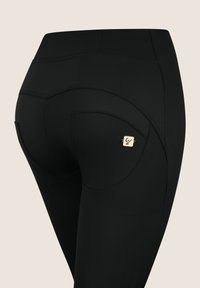 Mustat legginsit, joissa on muotoiltu design kaarevilla saumoilla, sivutasku ja pieni kultainen koristersymboli. Sujuva, joustava kangas.