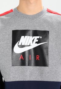 Grå sweatshirt med röda axelaccenter, prydd med en stor svart rektangulär logotyp med "NIKE" och "AIR" i vita och röda bokstäver.