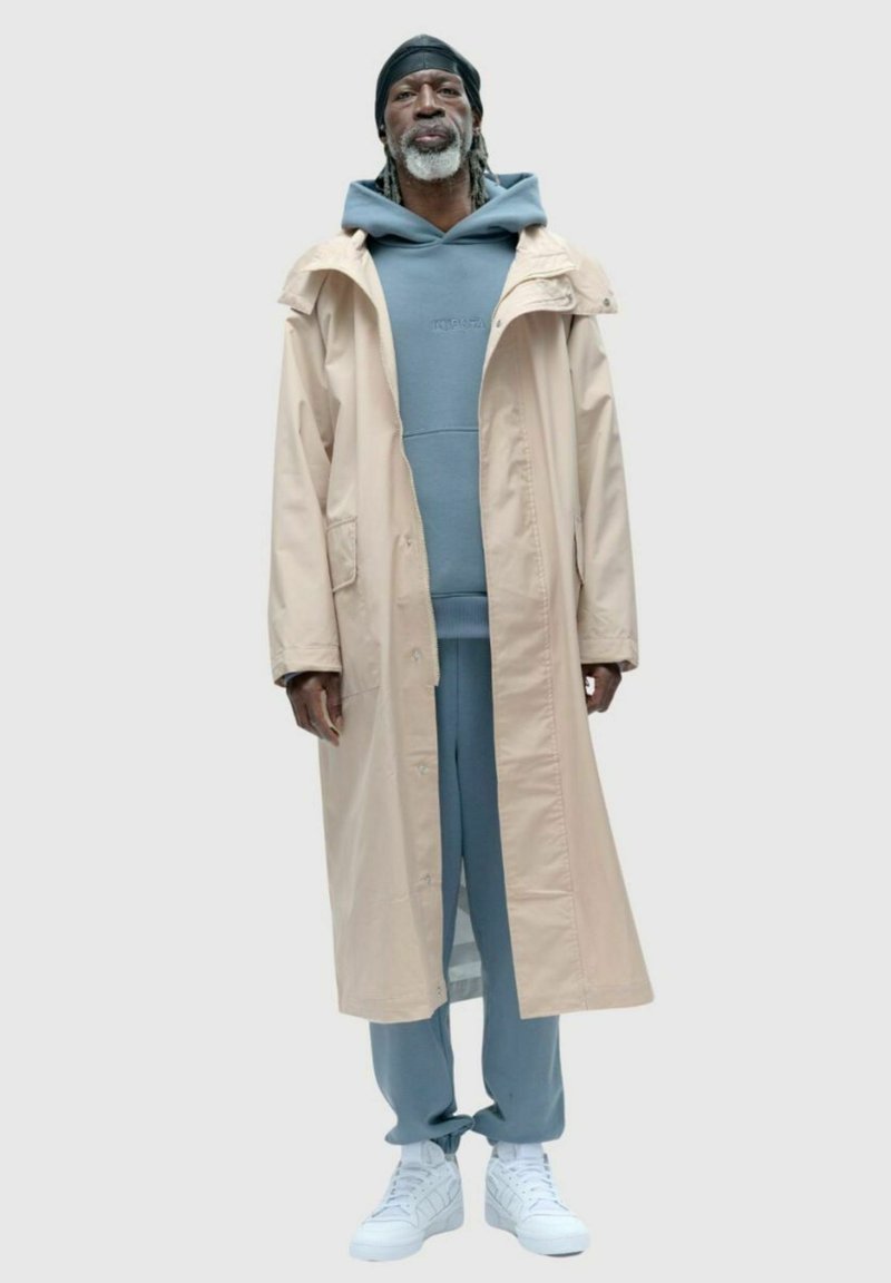 Beżowy długi płaszcz, niebieska bluza z kapturem, pasujące dresy, białe sneakersy. Płaszcz ma zapinanie na guziki i boczne kieszenie.