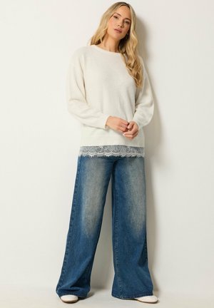 Femme aux longs cheveux blonds portant un pull blanc avec bordure en dentelle et un jean bleu à jambes larges, debout contre un mur clair uni.