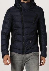 Mörkblå vadderad jacka med huva, med kviltad design, framdragkedjor och en sidoficka med dragkedja. Tillverkad av ett slätt, glänsande material.