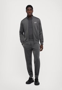 Joven vestido con un chándal EA7 gris oscuro y zapatillas negras, de pie con las manos en los bolsillos frente a un fondo neutro.