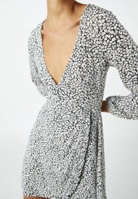 Vestido envolvente floral con mangas largas, que presenta un escote en V profundo. Fondo negro con margaritas blancas y rosas. Tejido ligero, textura suave.