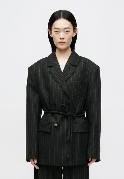 Blazer noir à rayures avec un design croisé, larges revers, une ceinture à nouer et deux poches avant. Tissu texturé avec des épaules structurées.