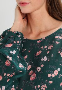 Groene bloemenblouse met een losse pasvorm, versierd met roze en witte bloemen, en een subtiele textuur. Bevat een knoopdetail op de manchette.