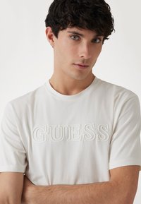 Camiseta de algodón blanca con el logotipo "GUESS" en relieve en la parte frontal, cuello redondo y mangas cortas con una textura suave.