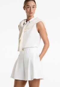 White sleeveless top with a collar, paired with a pleated white skirt. A light beige knitted sweater is draped over the shoulders.