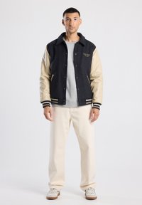 Black wool-blend varsity jacket with beige leather sleeves, grey ribbed cuffs, snap buttons, and side pockets; paired with light-colored pants.