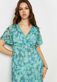 Robe turquoise avec imprimé léopard, présentant des manches volantées et un décolleté en V profond. Le matériau a une apparence douce et texturée.