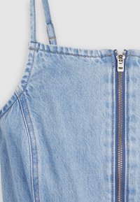 Vêtement en denim bleu clair avec fines bretelles et une fermeture éclair argentée centrale sur un fond uni.