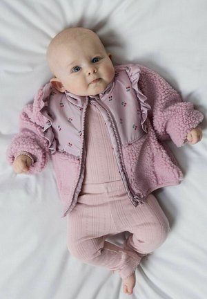 Bébé allongé sur une surface blanche, portant une veste rose texturée à imprimé floral et une tenue tricotée rose, regardant vers le haut avec une expression calme.