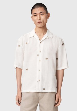 Jonge man draagt een wit overhemd met korte mouwen met kleine tijgergezichtprint, gecombineerd met lichtbeige broek, staand tegen een effen achtergrond.