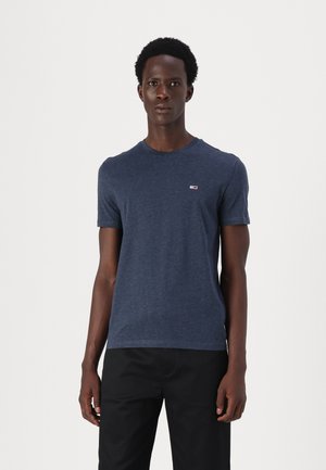 Uomo in piedi di fronte che indossa una t-shirt aderente blu navy a maniche corte con un piccolo logo sul petto e pantaloni neri su uno sfondo semplice.