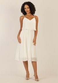 Weißes, plissiertes Kleid mit V-Ausschnitt und dünnen Trägern, das einen geschichteten Oberteil und einen fließenden Rock hat, der unter das Knie fällt.