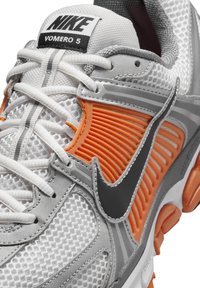 Nike Vomero 5 running shoe featuring a white mesh upper, orange ribbed accents, gray overlays, and a black logo. Textured sole with cushioning.