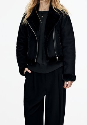 Woman wearing a black faux fur-lined biker jacket over a black knit sweater and loose black trousers, standing with one hand in pocket.