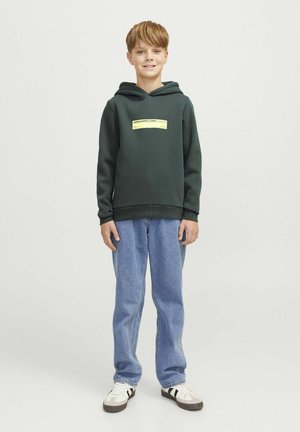 Felpa verde scuro con tasca a marsupio e etichetta grafica frontale, abbinata a jeans azzurri dalla vestibilità comoda e sneakers bianche.