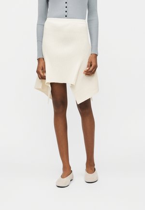 Femme portant une jupe asymétrique crème côtelée, un haut gris à manches longues boutonné, et des chaussures crème sans lacets, debout devant un fond blanc.