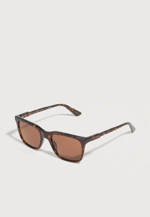Lunettes de soleil rectangulaires en écaille de tortue avec des verres marron et des branches fines posées sur une surface blanche.