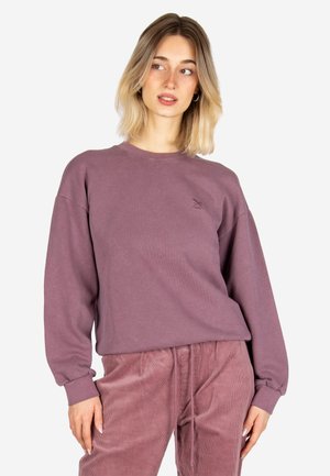 Junge Frau mit schulterlangen blonden Haaren, die einen mauvefarbenen Pullover und passende Cordhosen trägt und nach links schaut.