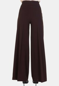 Pantaloni scuri marroni a vita alta e gamba larga indossati con scarpe con tacco nere, mostrati di spalle su uno sfondo bianco semplice.