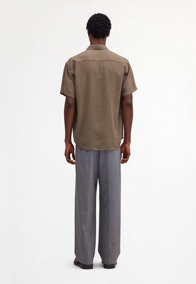 Personne debout, de dos, portant une chemise marron à manches courtes et un pantalon gris ample, sur un fond clair uni.
