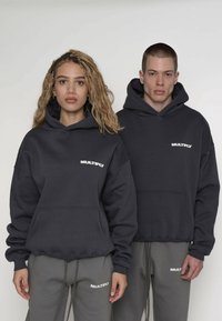 Hanorace gri închis cu buzunar frontal și logo-ul „MULTIPLY” alb. Asortate cu pantaloni de trening gri cu același logo.