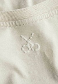 Lichtbeige stof met een gladde textuur, voorzien van een geborduurd scharenontwerp in een iets verhoogde steek nabij de halslijn.