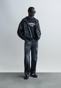 Fekete bőrbombázó dzseki fekete grafikás póló fölött, kopott kék farmernadrággal és fekete sportcipővel párosítva. Nagyméretű napszemüveg teszi teljessé a megjelenést.