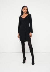 Sieviete ar cirtainiem matiem, valkājot melnu pogu apģērbu ar plata apkakli, melnas zeķubikses un melnus augstpapēžu apavus, stāvot pret balto fonu.