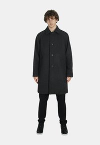 Cappotto di lana nera con un motivo texturizzato, chiusura frontale con bottoni e colletto classico, abbinato a pantaloni neri slim e sneakers nere.