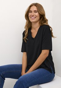 Čierne tričko s výstrihom do V, krátkymi rukávmi a voľným strihom, so bočnými rozparkami; skombinované s modrými džínsy. Model posedený na bielom pozadí.