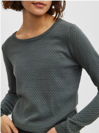 Ženska nosi temno sivo teksturirano dolgo-sleeve pulover z subtilnim vzorcem satovja, roke so ji sproščene ob pasu.