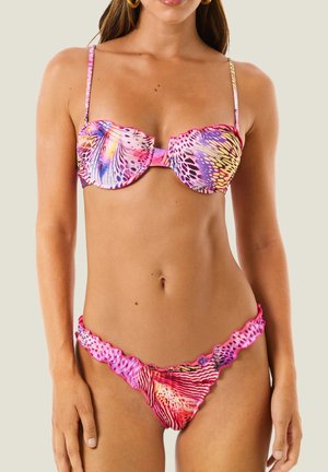 Donna che indossa un bikini colorato e con fantasia, con bordi arricciati e sottili spalline, in piedi davanti a uno sfondo chiaro e uniforme.