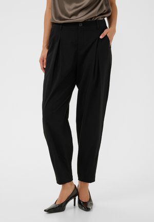 Femme portant un pantalon noir plissé et fuselé avec des talons hauts noirs pointus, une main dans la poche et un haut en satin marron.
