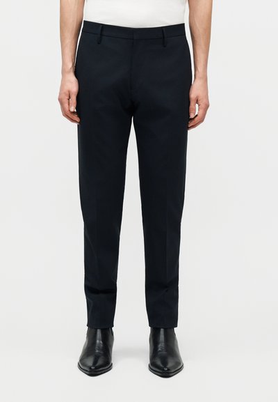 Άνδρας που φοράει στενά μαύρα παντελόνια φόρμας και μαύρα δερμάτινα μποτάκια αστραγάλου, στέκεται μπροστά σε απλό λευκό φόντο.