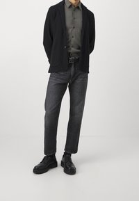Schwarze strukturierte Jacke, grau knöpfbares Hemd und dunkle Denim-Jeans, kombiniert mit schwarzen Schnürschuhen. Einfaches Design, neutrale Farbpalette.