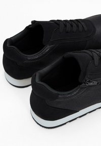 Scarpe da ginnastica nere realizzate con un mix di materiali lisci e testurizzati, dotate di un design con lacci, dettaglio con zip e suola bianca a contrasto.