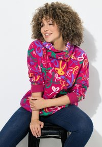 Felpa fucsia con motivi floreali in verde, viola e bianco. Presenta un collo avvolgente e polsini a coste. Indossata con jeans scuri.