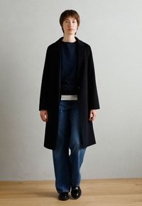 Manteau en laine noir sur un pull bleu marine, associé à un jean bleu à jambes larges. Texture lisse, fermeture boutonnée et silhouette droite.