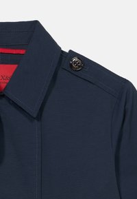 Γιακάς πουκάμισου σε ναυτικό μπλε με επίθετες επιγονατίδες στον ώμο, στερεωμένες με μαύρο κουμπί, με κόκκινο εσωτερικό ύφασμα ορατό στη γραμμή του λαιμού.