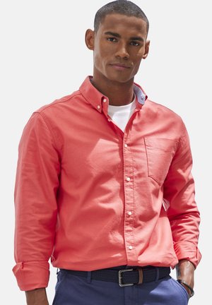 Korallfarbenes Button-up-Hemd mit Brusttasche, hochgekrempelten Ärmeln und einem kontrastierenden Innenkragenmuster. Wird über einem weißen Hemd getragen.