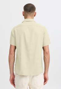 Chemise à manches courtes beige clair en tissu texturé, dotée d'un col classique et d'un ourlet arrondi. Détails de couture visibles.